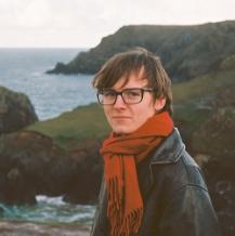 Student with dark framed glasses and red scarf with the sea and cliffs in the background.